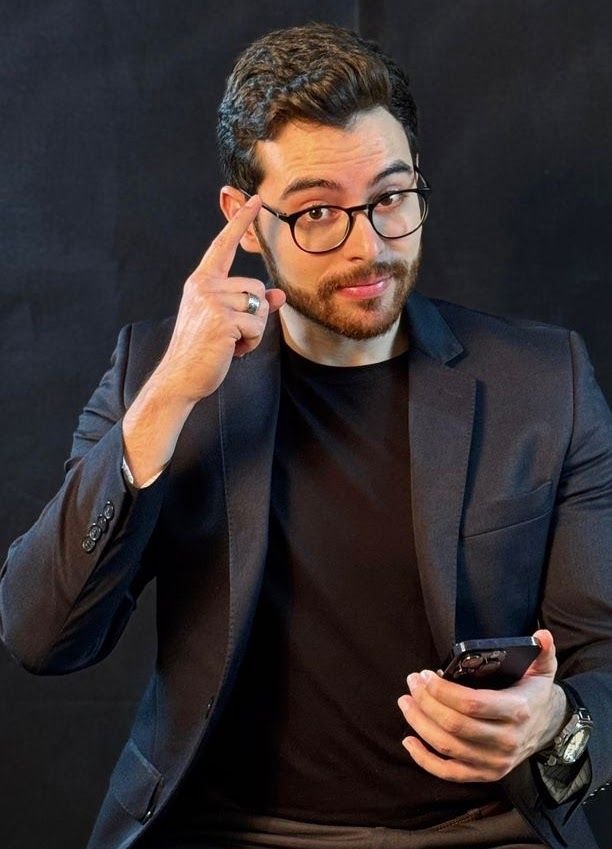 Retrato do Barbosa de óculos e blazer escuro, com o indicador na têmpora e o celular na mão